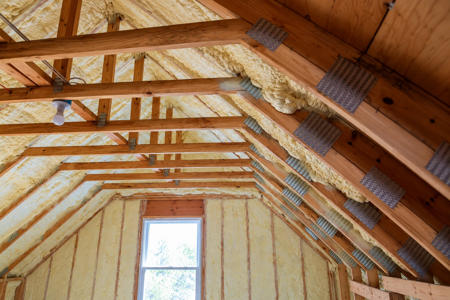 Attic framing with yellow-white spray foam on the roof and walls, exposed wooden trusses, and a window.