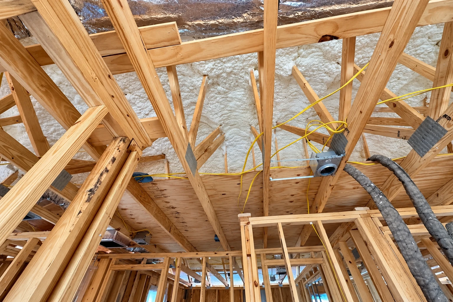 Exposed wooden roof framing is filled with white spray foam insulation, with ducts and yellow wiring running through the structure.