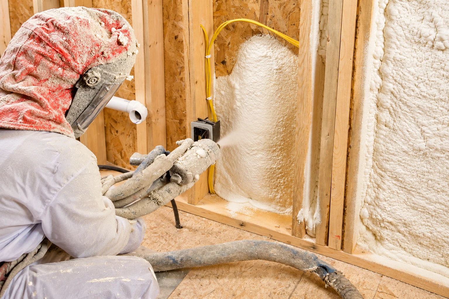 A worker in protective gear sprays expanding foam insulation into a wood-framed wall cavity.